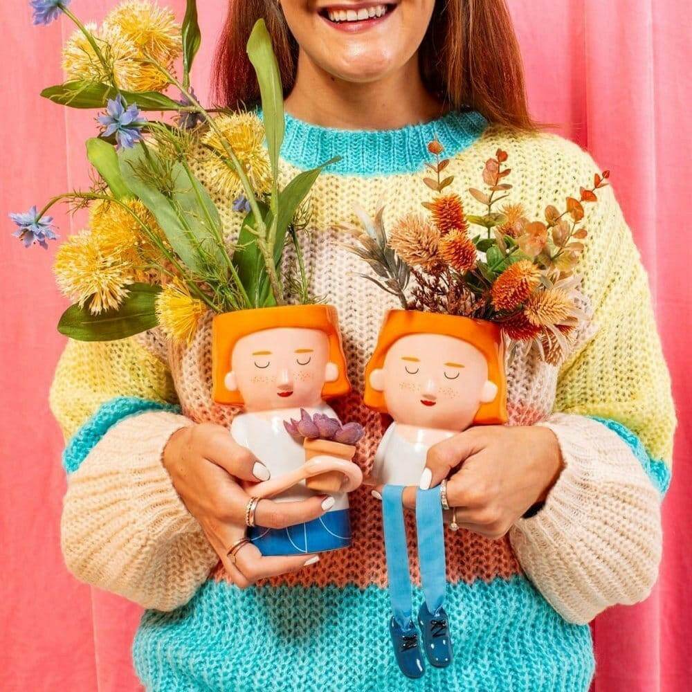 Orange hat plant pot figure with dangling legs and blue shoes, holding a floral bouquet
