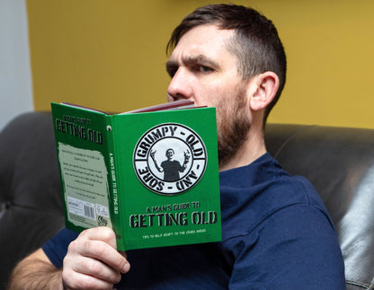 Man sitting on a couch reading a green “Getting Old” guide book